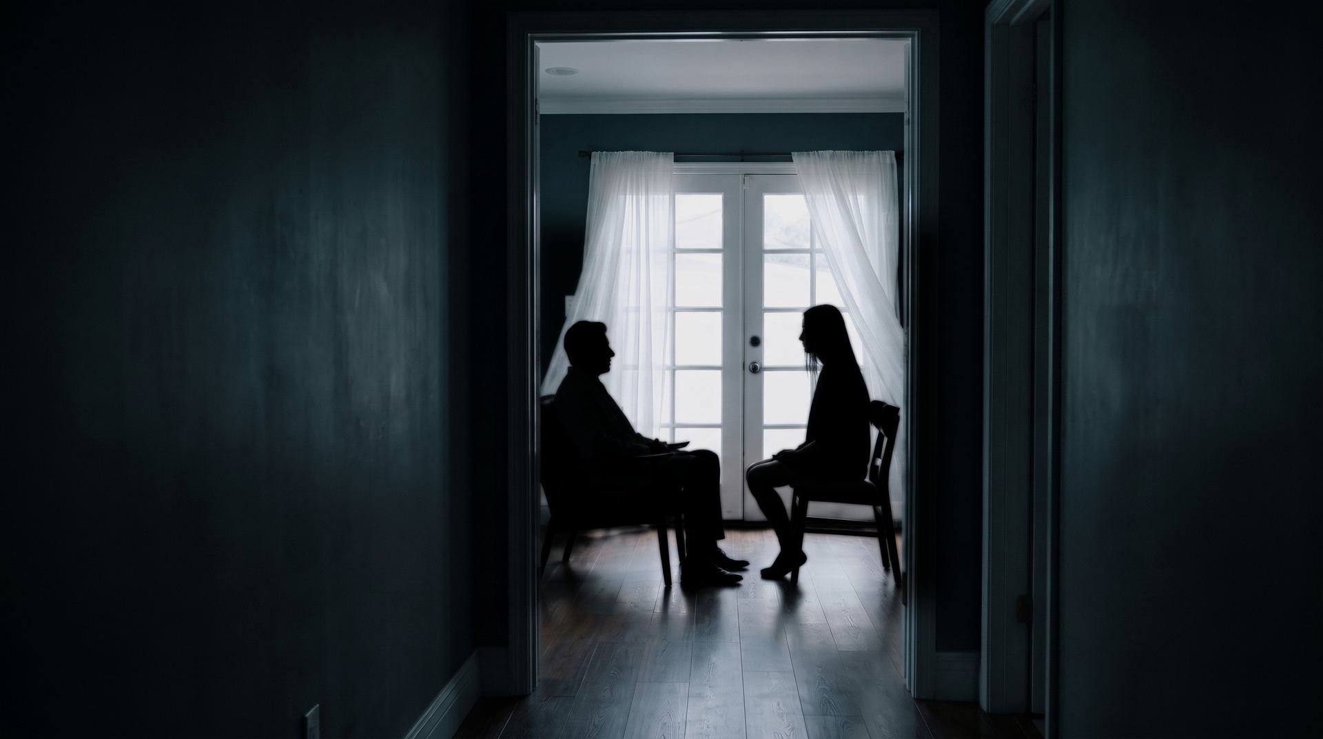 At the end of a dark hallway, through an open doorway, two figures sit facing each other in silhouette against a curtained window.