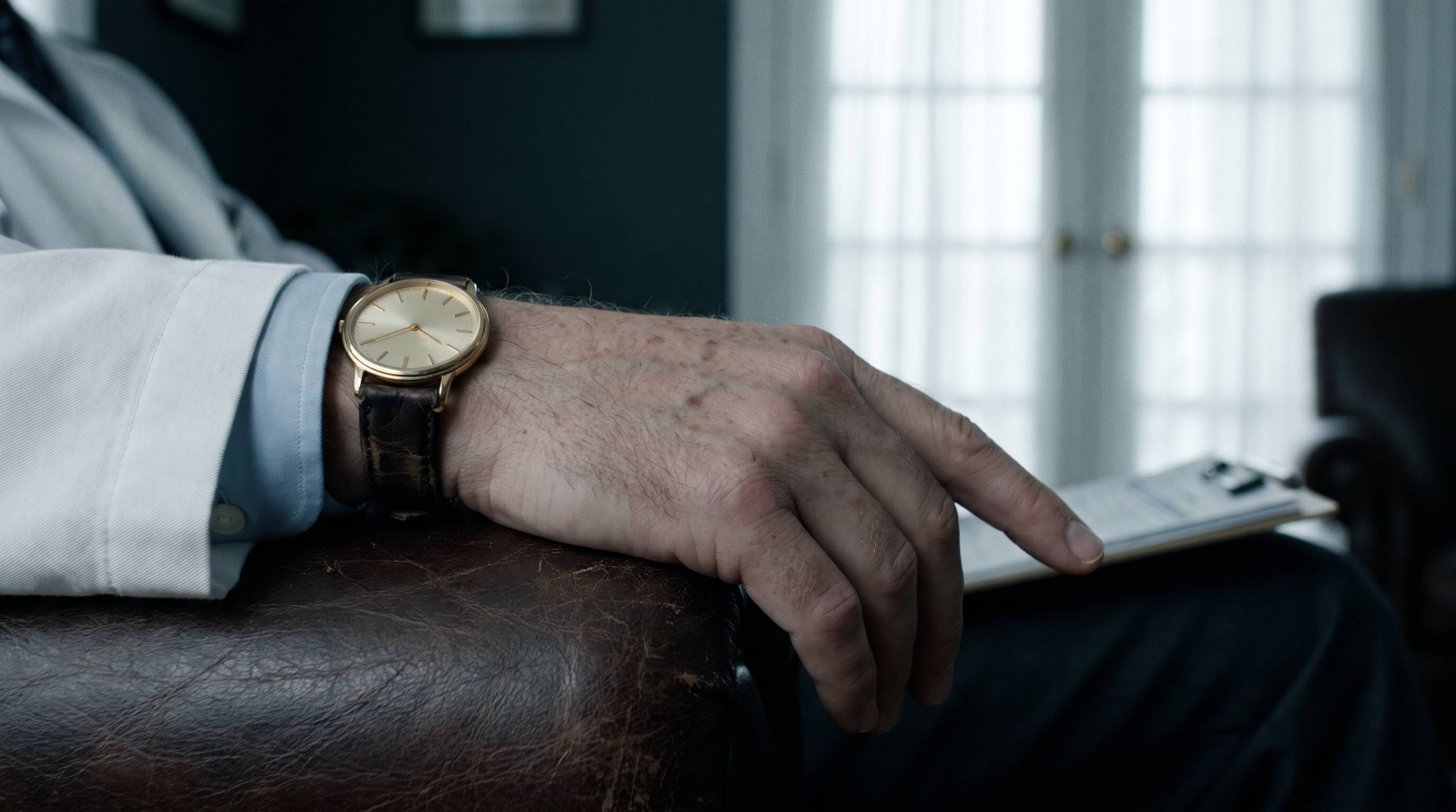 A gold dress watch on a man’s wrist, resting on the arm of a leather chair.